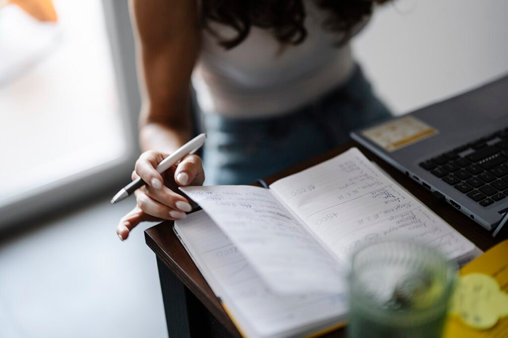 Pessoa sentada à mesa fazendo anotações em um caderno, ao lado de um notebook